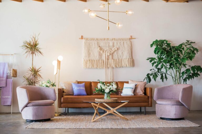 Brown leather sofa with pillows, two pink chairs, macrame wall hanging, and plants.