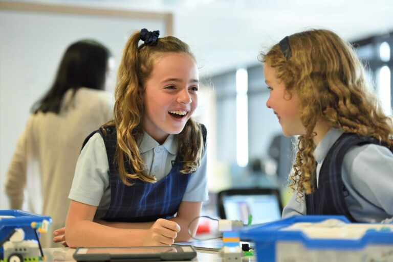 Two schoolgirls in uniform laugh while working on a STEM project with building blocks and a tablet.